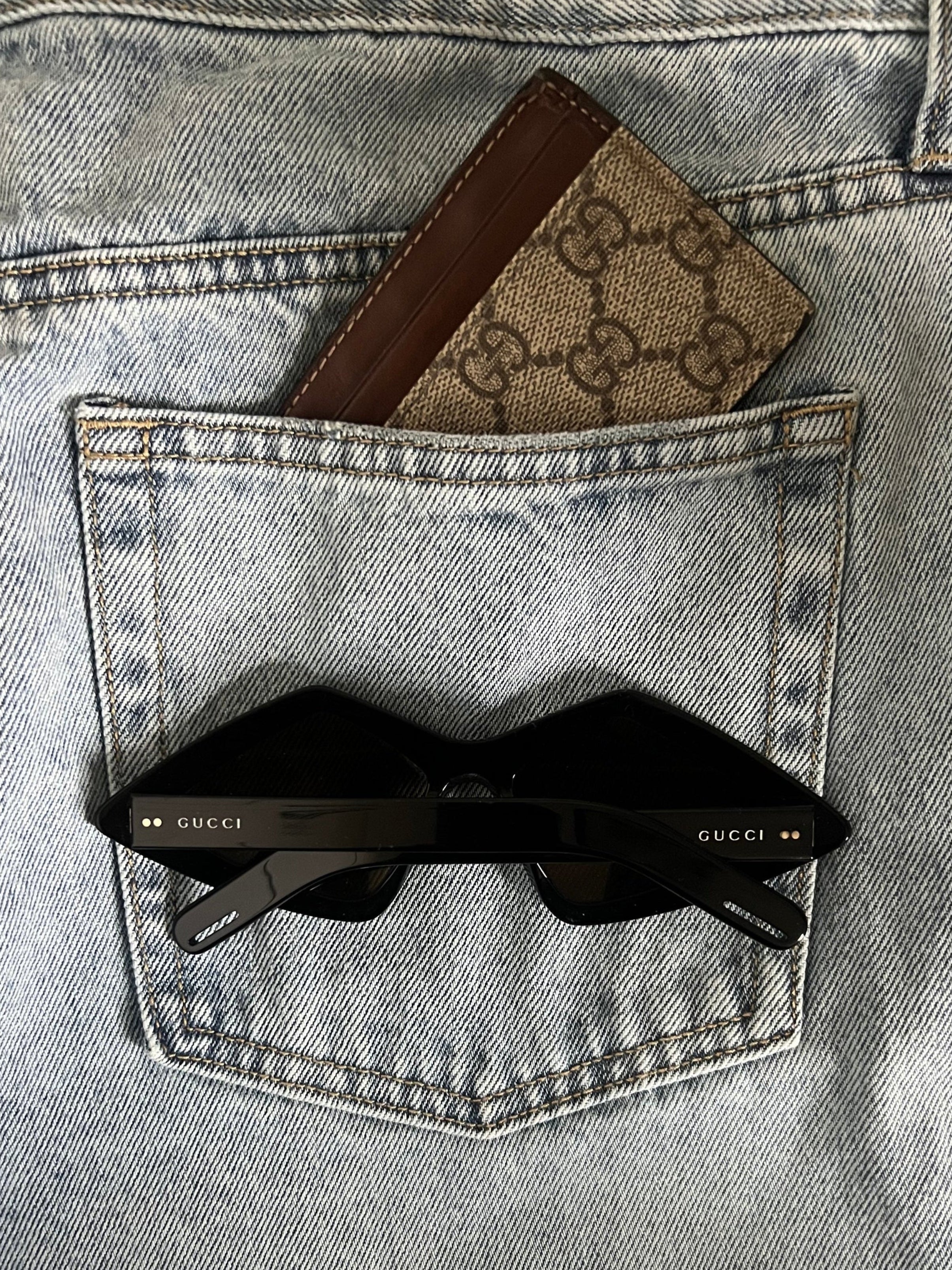 Black Gucci sunglasses in a pocket with a brown wallet on light blue denim jeans.